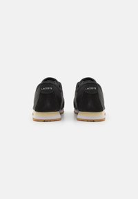 Baskets en suede noires avec des accents blancs, présentant un bout arrondi, une semelle rembourrée et un logo Lacoste bien visible au niveau du talon.