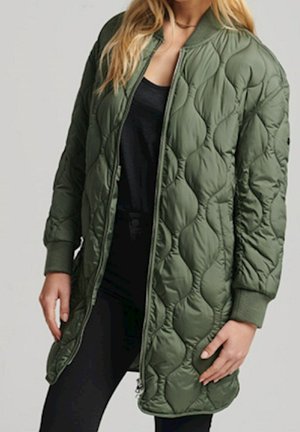 Une femme porte une longue veste bomber matelassée verte olive sur un top noir et un legging, debout avec les mains détendues sur les côtés.