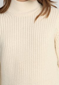 Cremfarbener, grob gestrickter Rollkragenspullover, getragen, der das strukturierte Muster und einen Teil der braunen Haare einer Person zeigt.