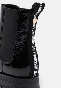 Bottine en cuir verni noir avec panneau latéral élastique, languette de tirage brandée portant l'inscription "LOVE MOSCHINO" et petit pendentif en forme de cœur doré.