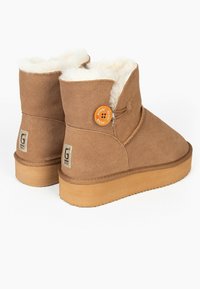 Hnědé semišové kotníkové boty s huňatou bílou podšívkou, kulatým oranžovým knoflíkem a světle hnědou gumovou podrážkou. Mají logo na patě.