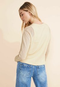 Strickpullover in hellem Beige mit einem strukturierten Netzdesign, kombiniert mit blauen Jeans. Hat einen Rundhalsausschnitt und lange Ärmel.