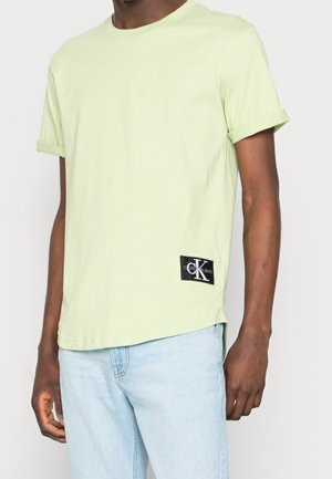 Mann trägt ein hellgrünes T-Shirt mit kurzen Ärmeln und einem schwarzen Calvin Klein Jeans Logo-Patch, kombiniert mit hellblauen Jeans.