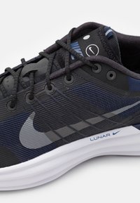 Fekete és sötétkék sportcipő textúrázott szövet felsőrészével, megerősített fűzőlyukakkal és fehér középtalppal. Szürke swoosh logóval és "LUNAR" felirattal rendelkezik.