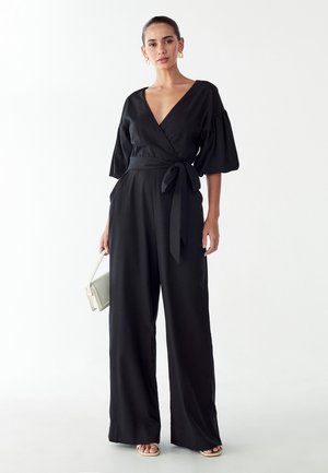 Combinaison noire avec un décolleté en V profond, des manches ballon et une taille nouée. Le tissu semble lisse, avec un pantalon à jambes larges. Accessoires minimalistes.