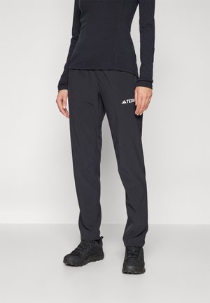 Personne portant un haut noir à manches longues, un pantalon adidas Terrex noir et des chaussures de sport noires, debout devant un fond clair uni.