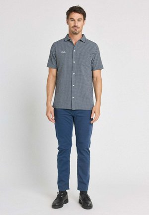 Homme debout portant une chemise à manches courtes à carreaux boutonnée, un pantalon bleu et des chaussures noires, sur un fond clair uni.