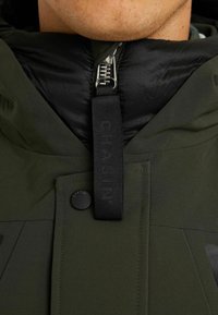 Kołnierz ciemnozielonej kurtki z czarnym zamkiem błyskawicznym i materiałową metką. Charakteryzuje się matową teksturą i ciemnymi panelami akcentowymi. Widoczny detal zatrzasku.