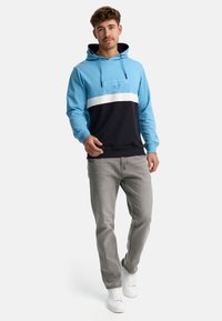 Hoodie coloré en bleu et noir avec une rayure blanche, capuche avec cordon de serrage et poignets côtelés. Porté avec un jean gris clair et des baskets blanches.