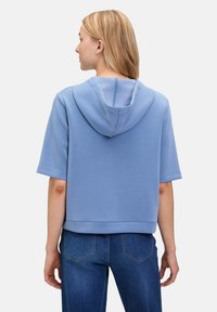Jasnoniebieska bluza z kapturem z krótkimi rękawami, luźny krój i szew centralny z tyłu. Połączona z dżinsami.