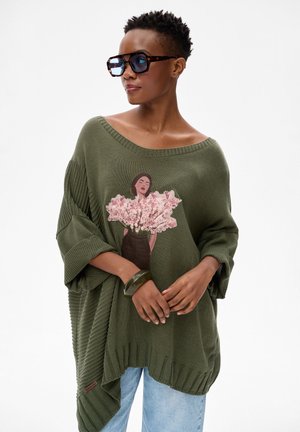 Kobieta w oversize'owym oliwkowozielonym swetrze z nadrukiem kwiatów i sylwetką kobiety, duże okulary przeciwsłoneczne, bransoletka i jasnoniebieskie dżinsy na białym tle.