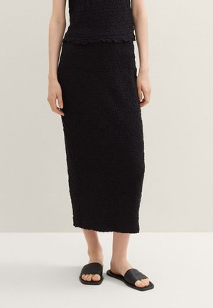 Schwarzes, strukturiertes zweiteiliges Outfit mit einem ärmellosen Oberteil und einem langen Rock, kombiniert mit flachen schwarzen Slipper-Sandalen.