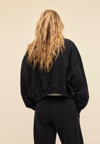 Persona bionda con capelli mossi che indossa una felpa nera larga e pantaloni, in piedi con le mani nelle tasche, di spalle alla fotocamera.