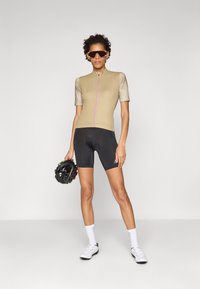 Beige sykkelskjorte med korte ermer, rosa glidelås, svarte sykkelshorts og hvite sykkelsko. Holder en svart hjelm.
