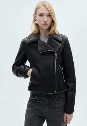 Černá kožená bunda s šedým kožešinovým límcem, zapínání na zip a bočními kapsami. Tvarované rukávy a zkrácený design. Nositá přes šedý top.