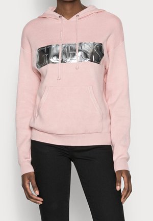 Personne portant un sweat à capuche rose clair avec un logo "GUESS" réfléchissant argenté sur la poitrine et un pantalon noir, les mains détendues le long du corps.