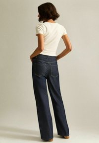 Donna con i capelli corti castani che indossa una maglietta bianca a maniche corte e jeans larghi di colore scuro, in piedi con le mani nelle tasche, rivolta di spalle.