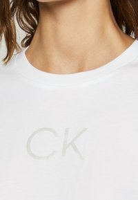 Bílý bavlněný tričko s jemnou texturou a světlé šedou tištěnou značkou "CK" na přední straně. Klasický design s kulatým výstřihem.