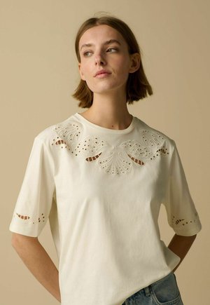 Jeune femme aux cheveux bruns courts portant une chemise blanche avec des motifs de papillons en ajouré sur la poitrine et les manches.
