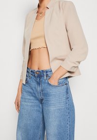 Chaqueta corta beige con mangas tres cuartos, combinada con un top beige ajustado y unos vaqueros azules sueltos, con botón y bolsillos frontales.