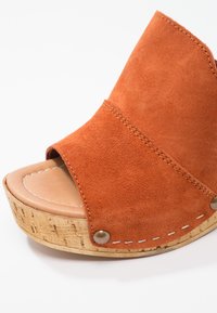 Sandale compensée en daim orange avec bout ouvert, plateforme en liège et détails de couture. Présente des bords arrondis et un décor en métal.