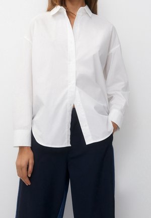 Personne portant une chemise blanche ample à manches longues boutonnée et un pantalon large bleu marine foncé, debout avec une main dans la poche.
