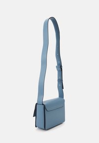 Obdélníková světle modrá crossbody taška s klopovým uzávěrem a nastavitelným širokým popruhem na bílém pozadí.