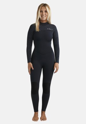 Młoda kobieta stojąca boso, ubrana w czarny, pełny kombinezon neoprenowy z wzmocnionymi nakolannikami i wzorzystymi rękawami, uśmiechająca się do aparatu.