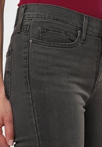 Vaqueros de mezclilla negros con una textura suave, diseño de cinco bolsillos y detalles de costura visibles, mostrados de lado con la colocación de la mano.