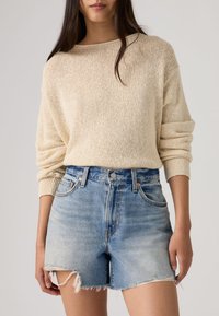 Γυναίκα που φοράει μπεζ πλεκτό πουλόβερ με γυρισμένα μανίκια και ψηλόμεσο ανοιχτό μπλε τζιν σορτς με φθαρμένα άκρα.