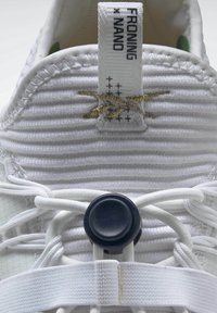 Gros plan sur la languette d'une basket blanche avec un tirant marqué Froning X Nano et un bloque-lacet circulaire noir sécurisant des lacets élastiques blancs.