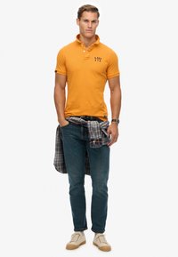 Orange polo-shirt med krave og tre knapper, kombineret med mørkeblå jeans og en ternet skjorte bundet om livet. Lyse sneakers på fødderne.