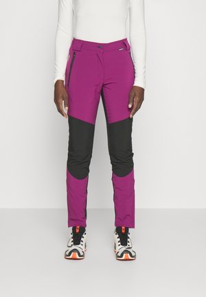 Person trägt zweifarbige lila-schwarze Outdoorhose mit Reißverschlusstaschen und weißen Sportschuhen mit orangefarbenen Akzenten.