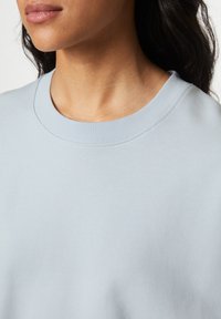 Primo piano di una persona che indossa una maglietta leggera blu con collo rotondo, con in vista il collo, le labbra e parte dei capelli scuri.