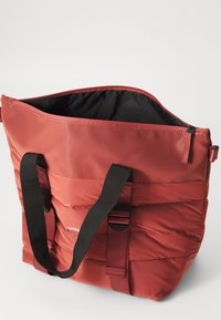 Rød, polstret totebag med en blank finish. Har svarte stoffremmer og en glidelås på toppen. Innvendig er den svart, noe som fremhever bagens tekstur.