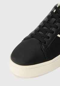 Mustast nahast sneaker ümmarguse varba, musta nööbiga disaini ja tekstuurse valge kummist tallaga. Küljel on kontrastne valge triip.