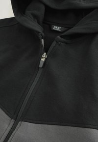 Haut à capuche zippé noir et gris avec une poche kangourou, un tissu texturé et un cordon de serrage, comportant une fermeture éclair visible et des détails de couture.