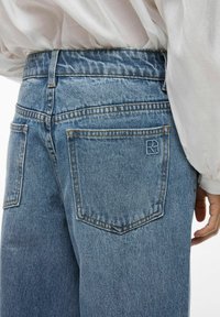 Jean en denim bleu clair avec une texture légèrement délavée, comprenant deux poches arrière et des détails cousus. La poche droite porte un logo.