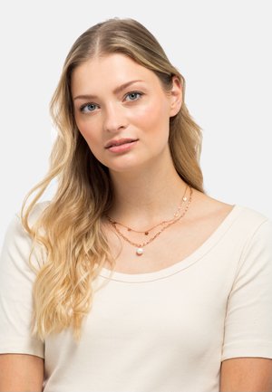 Collana a strati con catene in oro rosa, accentate da elementi circolari e texturizzati, e un grande pendente a forma di perla. Il modello indossa un top beige chiaro.