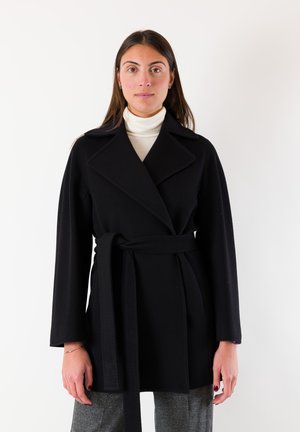 Cappotto nero avvolgente con colletto a rever, maniche lunghe e cintura frontale annodata. Realizzato in tessuto strutturato. Indossato sopra un dolcevita bianco.