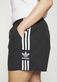 Pantaloni sportivi neri realizzati in tessuto leggero, con tre strisce bianche sui lati e un logo Adidas ricamato in bianco.