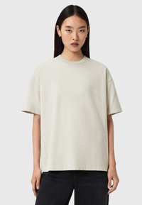 Φαρδιά ανοιχτό μπεζ κοντομάνικη μπλούζα από απαλό ύφασμα, με στρογγυλή λαιμόκοψη και κοντά μανίκια, με διακριτική λεπτομέρεια λογότυπου στο μπροστινό μέρος.