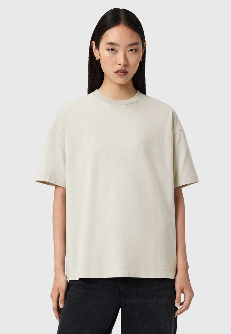 Φαρδιά ανοιχτό μπεζ κοντομάνικη μπλούζα από απαλό ύφασμα, με στρογγυλή λαιμόκοψη και κοντά μανίκια, με διακριτική λεπτομέρεια λογότυπου στο μπροστινό μέρος.