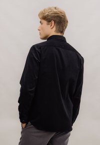 Svart kordfløyelskjorte med krage, lange ermer og knapplukking ved håndleddene. Har en jevn tekstur og en litt buet nederkant.