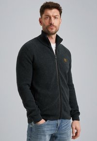 Zwarte ribgebreide zip-up trui met een hoge kraag, voorzien van een subtiele textuur en een klein vierkant logo op de borst.