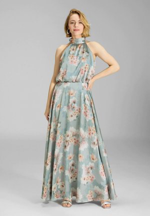 Femme portant une robe longue sans manches à col haut, bleu pastel avec un imprimé floral rose et blanc, posant avec une main sur la hanche.