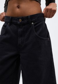 Osoba nosząca luźne czarne dżinsy, ciągnąca pas spodni na zewnątrz, aby pokazać nadmiar miejsca, widoczny brzuch i krótki ciemny top.