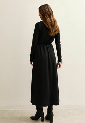 Mujer con cabello largo y castaño vistiendo un vestido largo negro y botas negras con tacón, de pie en el interior contra una pared lisa y clara.