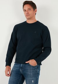 Tengerészkék pulóver hosszú ujjakkal, kerek nyakkivágással és raglán ujj kialakítással. Sima textúra, kis logóval a mellkason. Kék farmerrel viselve.