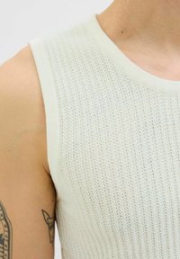 Licht beige mouwloos gebreid top met een semi-doorzichtig textuur en een ronde halslijn. Zichtbare stiksels langs de randen.